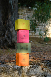 A stack of Huskke & Co sustainable candles in colorful packaging, balanced outdoors on a stone surface with a natural backdrop of trees and greenery.