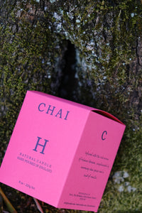 Huskke & Co Chai Candle packaging in vibrant pink, resting against textured tree bark. A subtle vanilla candle infused with warming chai spices.