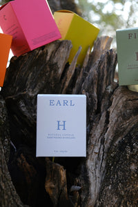 Huskke & Co Earl Candle in soft white packaging, nestled against rugged tree bark. A hand-poured lavender essential oil candle with a tranquil scent.